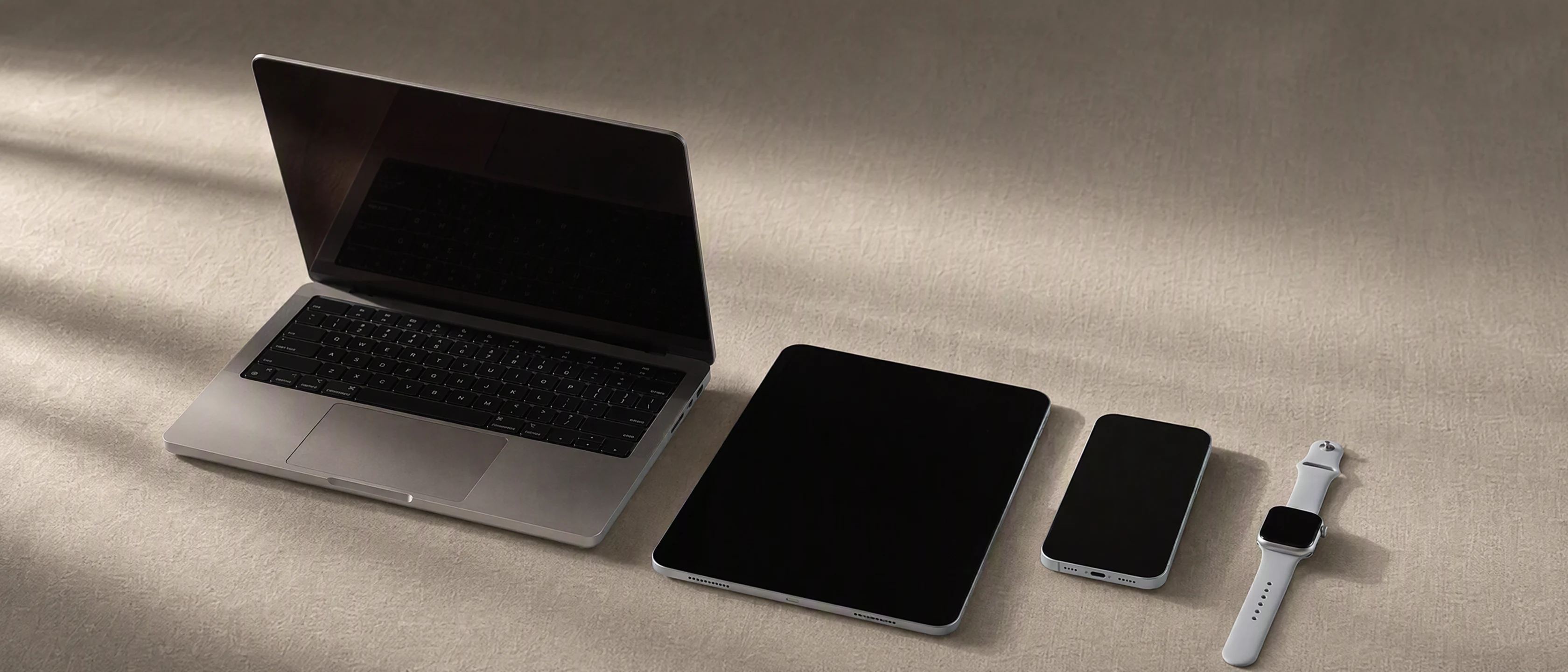 An iPhone, an Apple Watch, an iPad and a MacBook at rest together on a light linen surface in warm side light, their screens dark.
