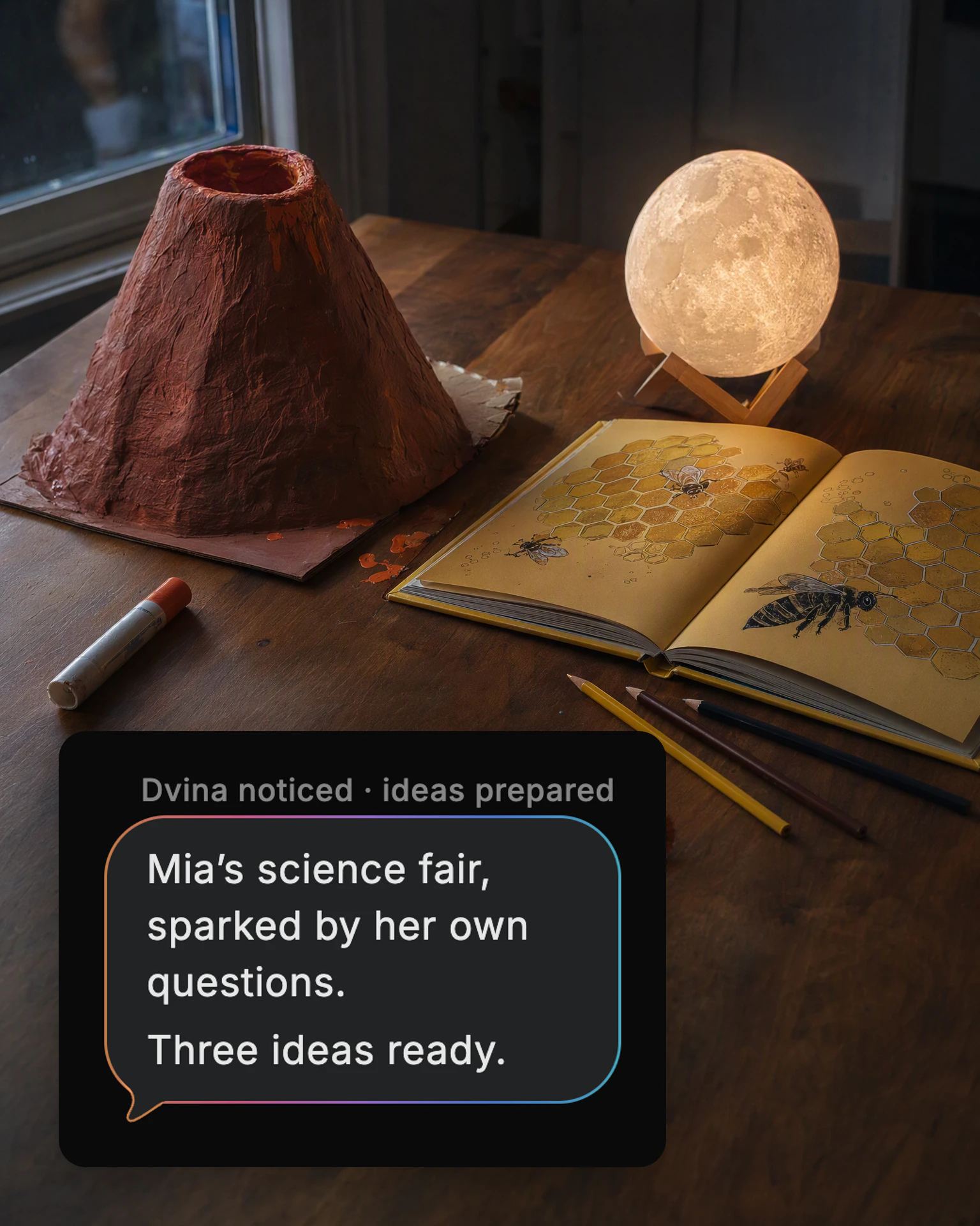 A kitchen table with a paper volcano, a book about bees and a moon lamp, and Dvina's card: “Mia’s science fair, sparked by her own questions. Three ideas ready.”