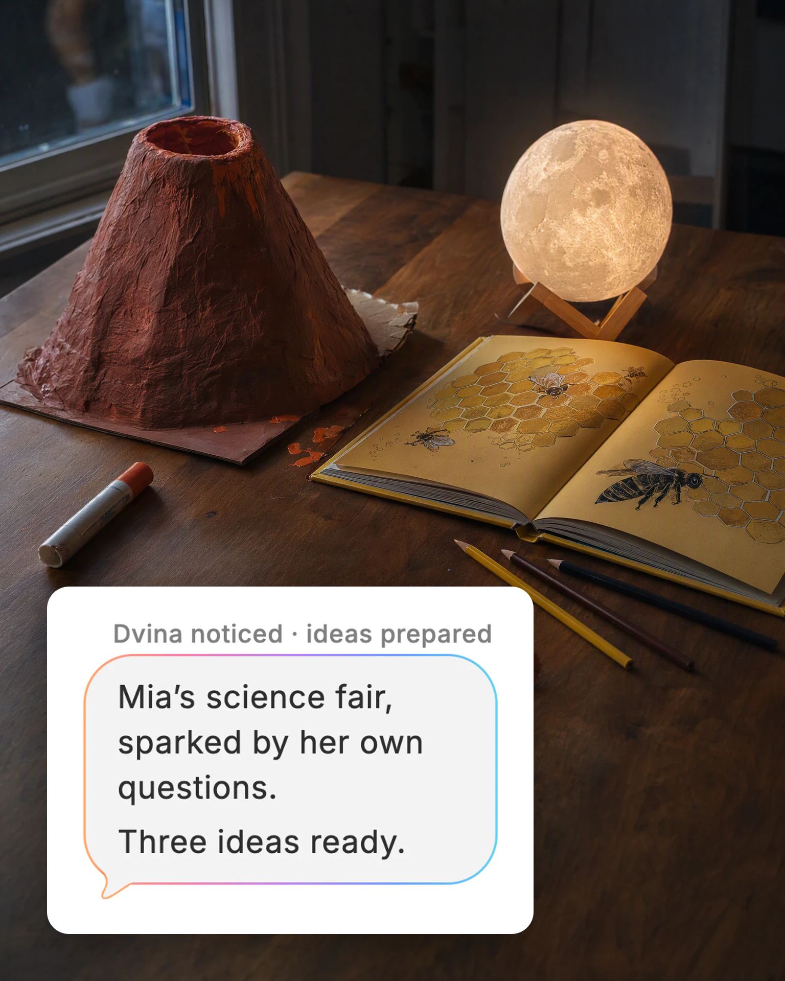 A kitchen table with a paper volcano, a book about bees and a moon lamp, and Dvina's card: “Mia’s science fair, sparked by her own questions. Three ideas ready.”