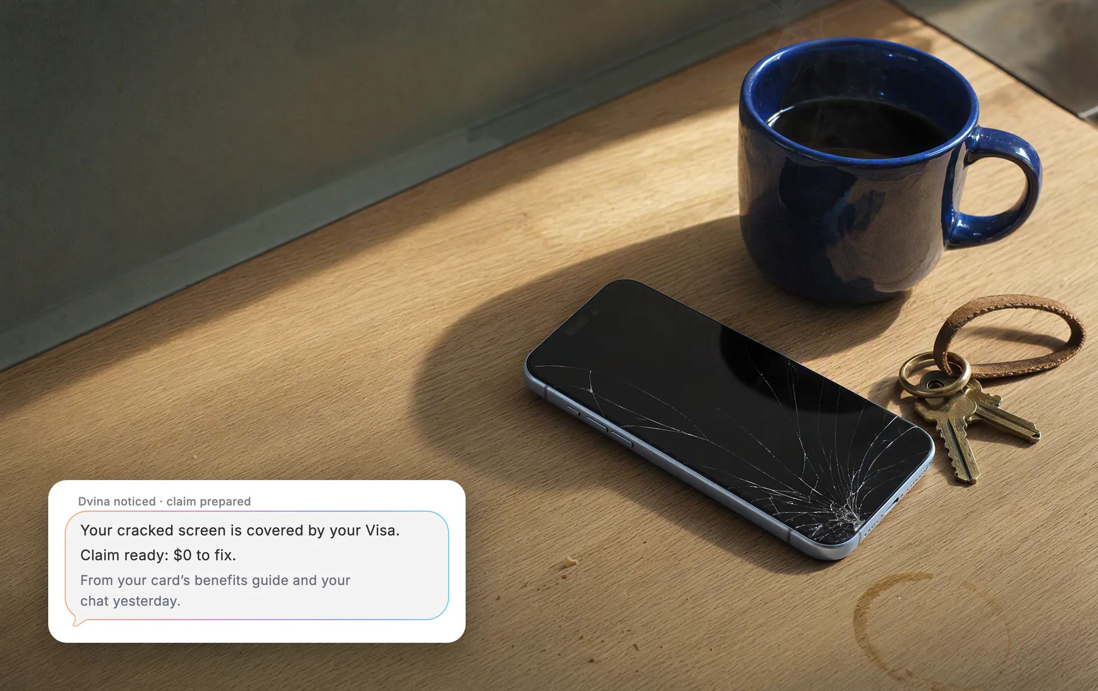 Morning light on a kitchen counter: a phone face-up with a cracked screen beside a cobalt coffee mug and keys, and Dvina's card, unasked: “Your cracked screen is covered by your Visa. Claim ready: $0 to fix.” From your card’s benefits guide and your chat yesterday.