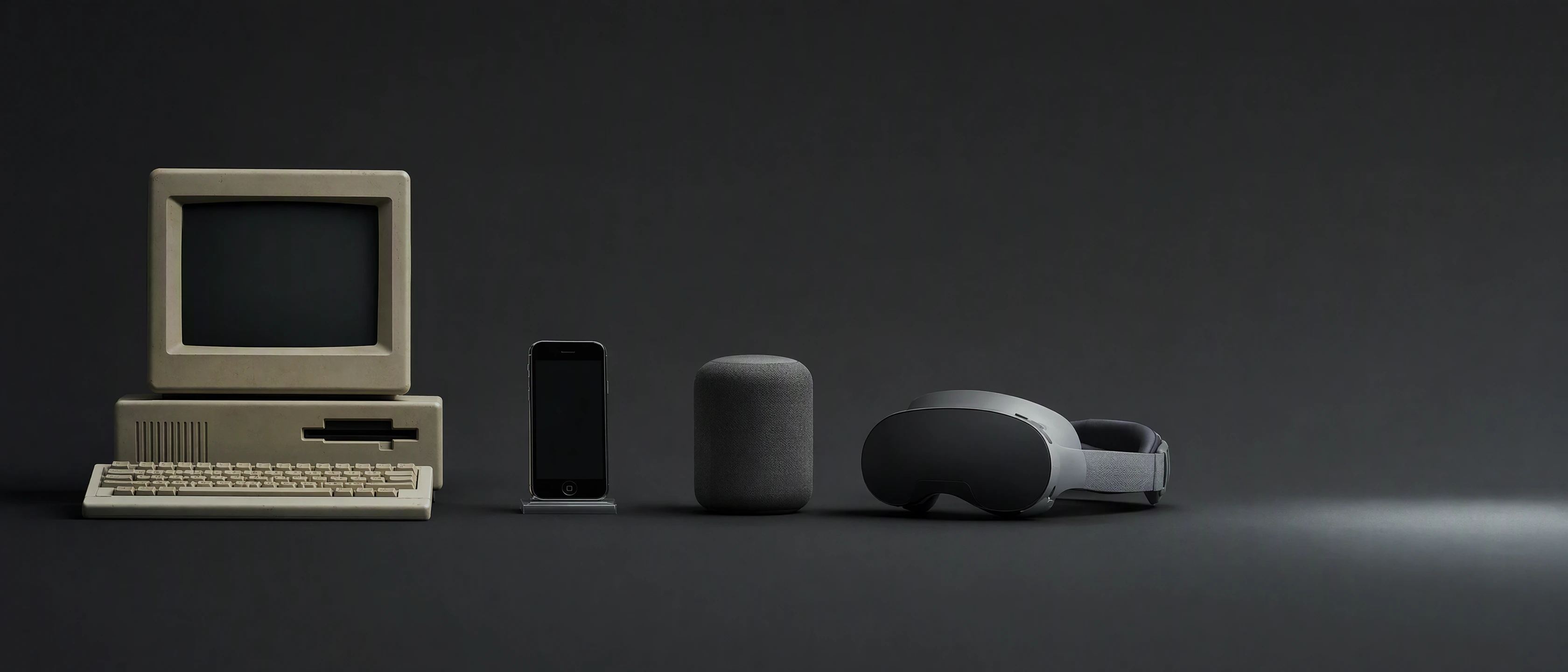 On one pale surface, in a line from left to right: a beige 1980s computer, a late-2000s smartphone, a fabric smart speaker, a mixed-reality headset, and then open space in soft light.
