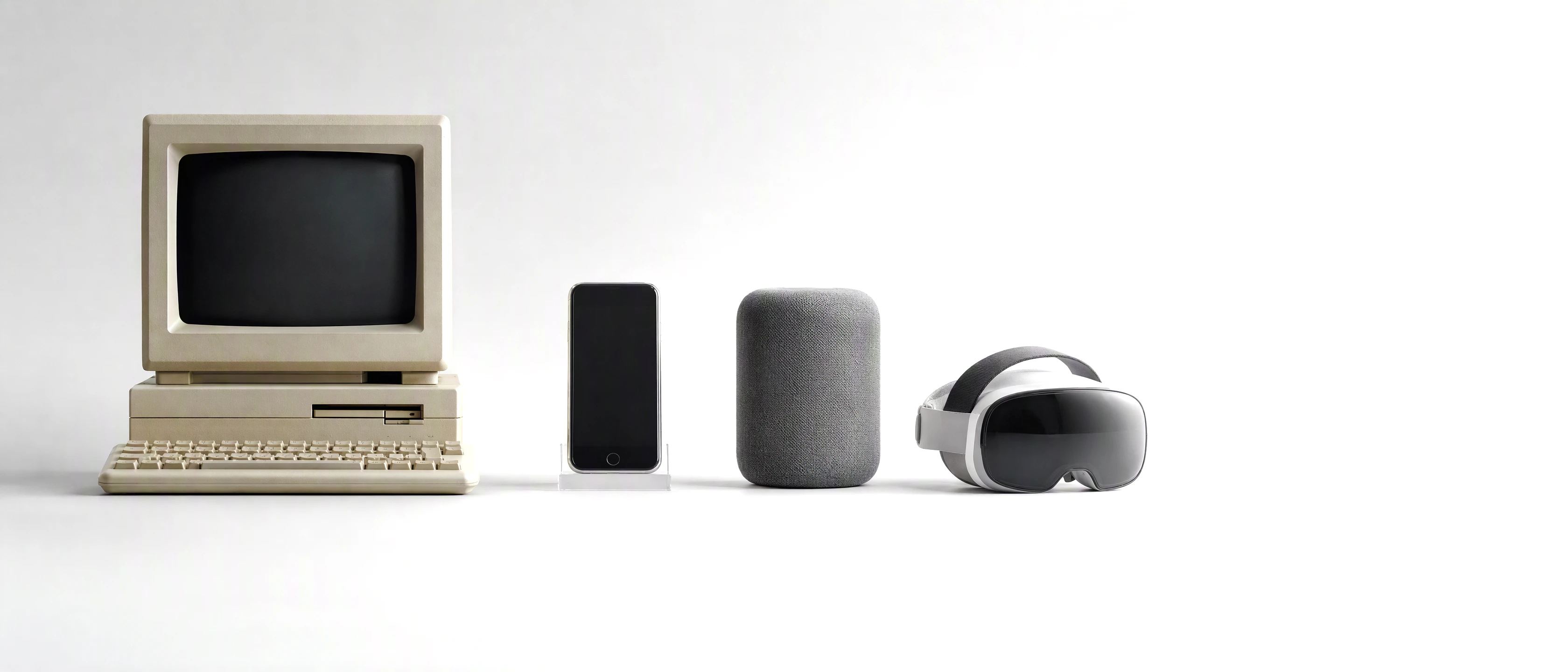 On one pale surface, in a line from left to right: a beige 1980s computer, a late-2000s smartphone, a fabric smart speaker, a mixed-reality headset, and then open space in soft light.