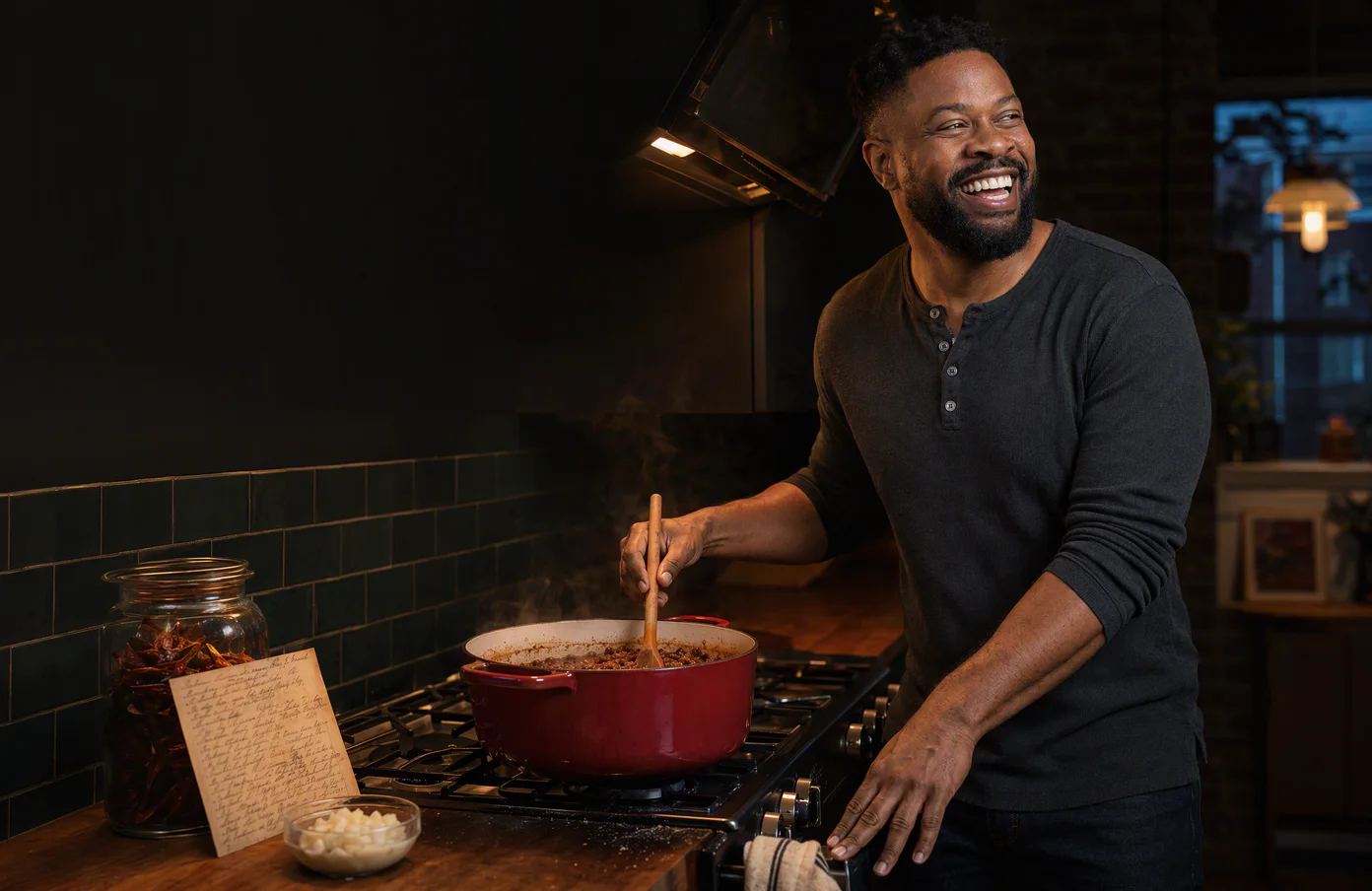 Marcus laughs at his stove on a Sunday evening, a wooden spoon in the pot, his grandmother's recipe card propped beside him.