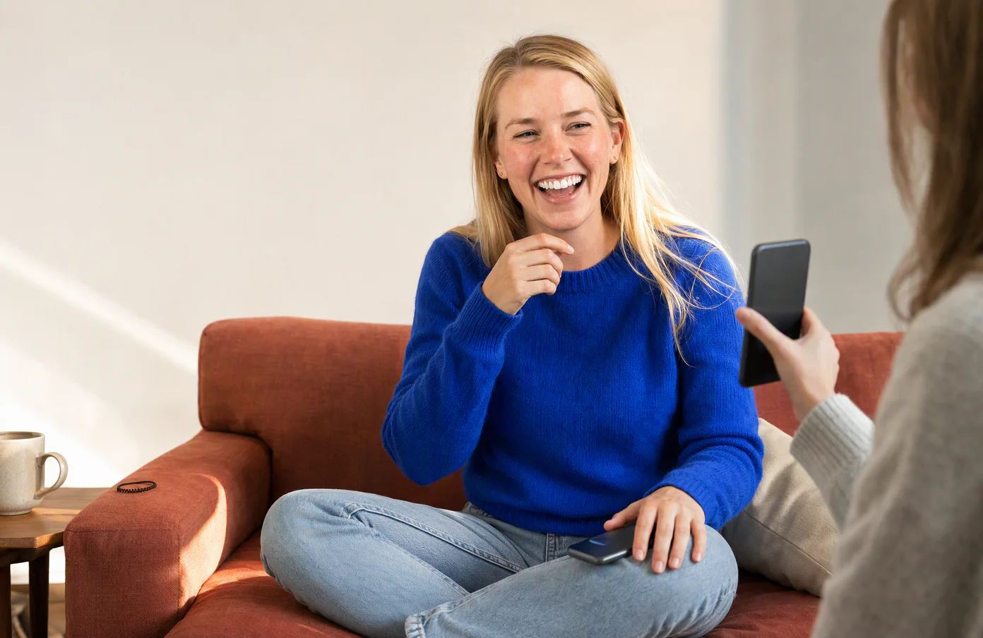 Emily laughs on a friend's couch as the friend shows her a photo, her own phone face down on her knee.