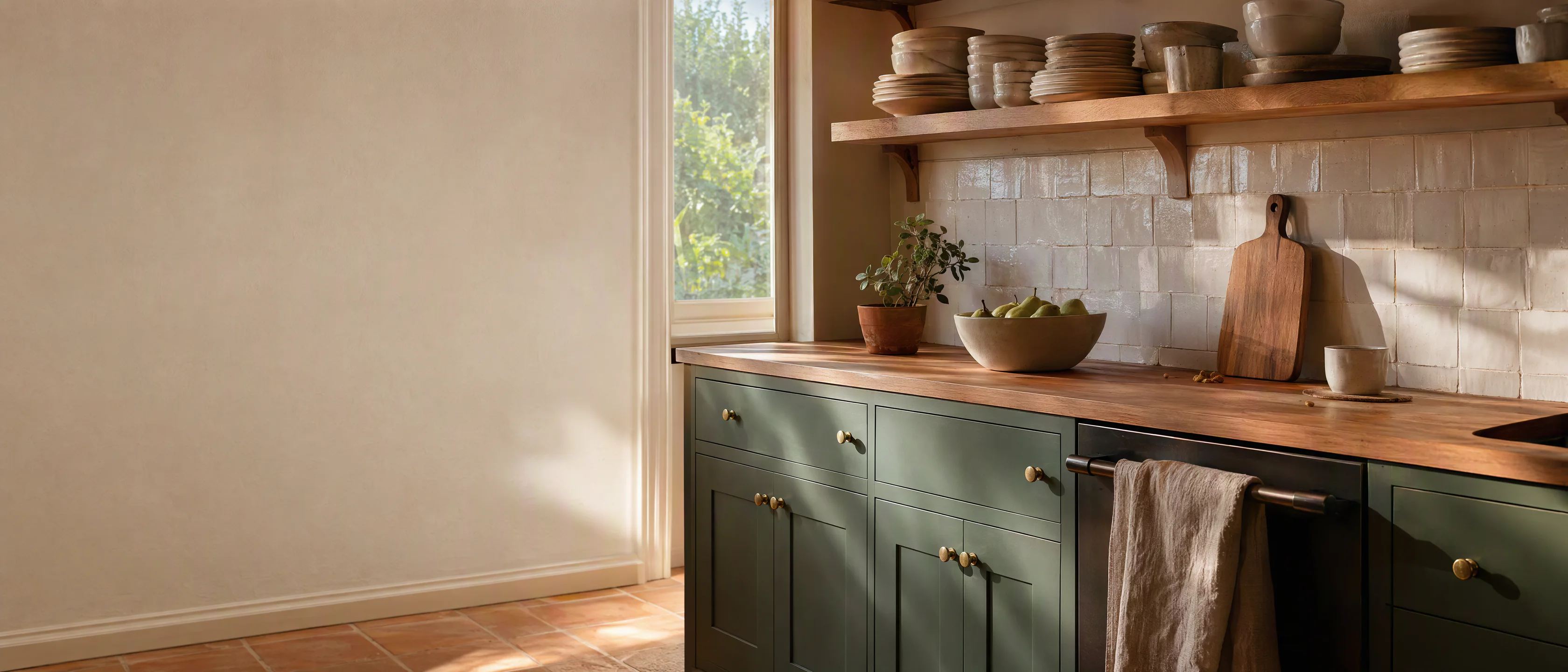 A finished kitchen in morning light: deep sage-green cabinets, open oak shelves with white ceramics and terracotta tile.