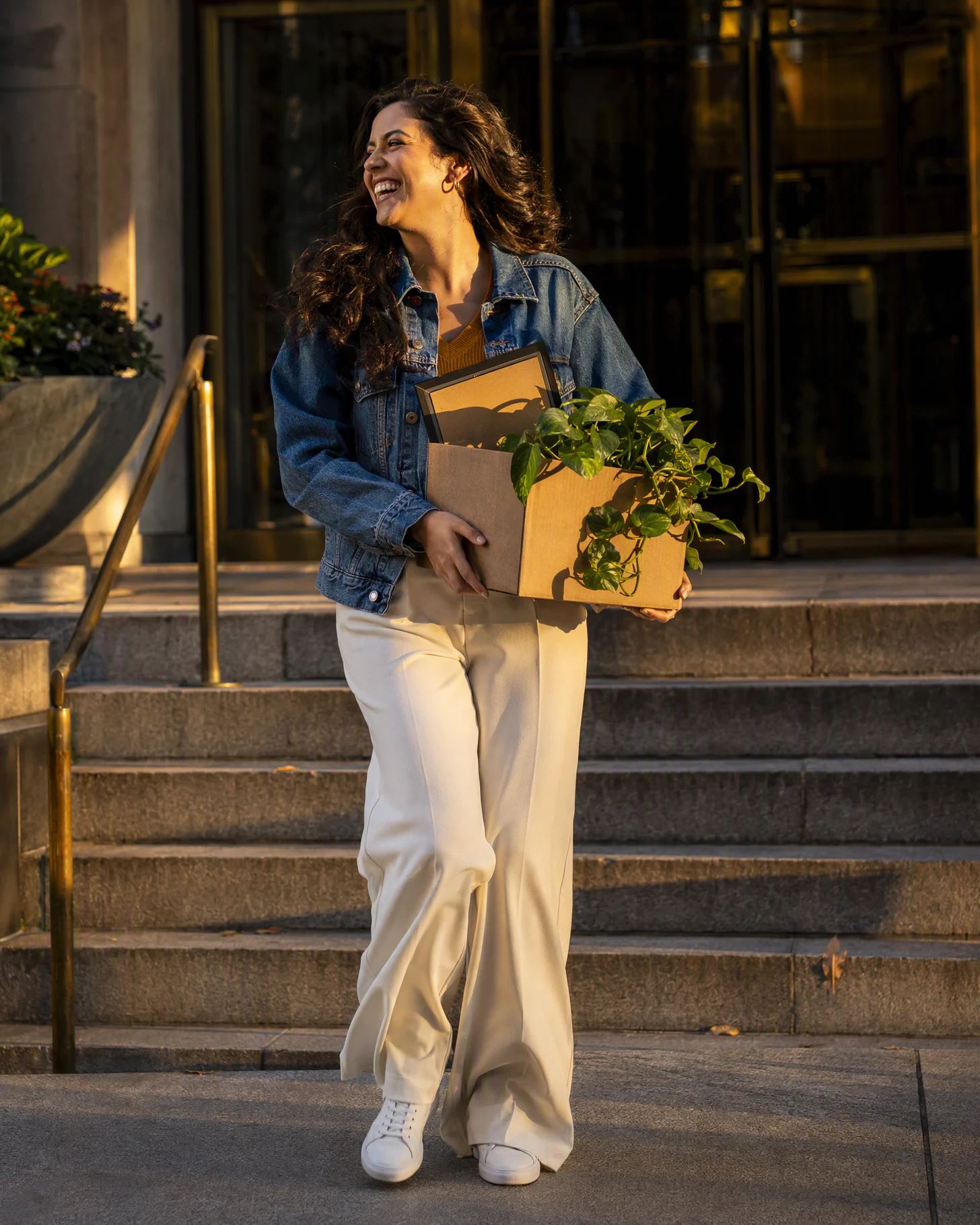 A woman laughing as she walks down the steps of her office at sunset, a box with a plant and a framed photo at her hip.