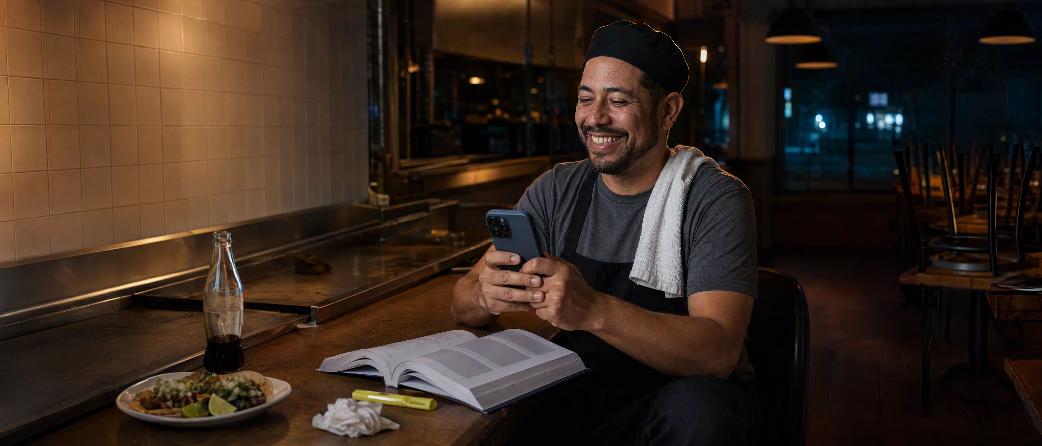 Juan on a counter stool in a closed taqueria after midnight, an accounting textbook beside a plate of tacos, grinning at his phone.