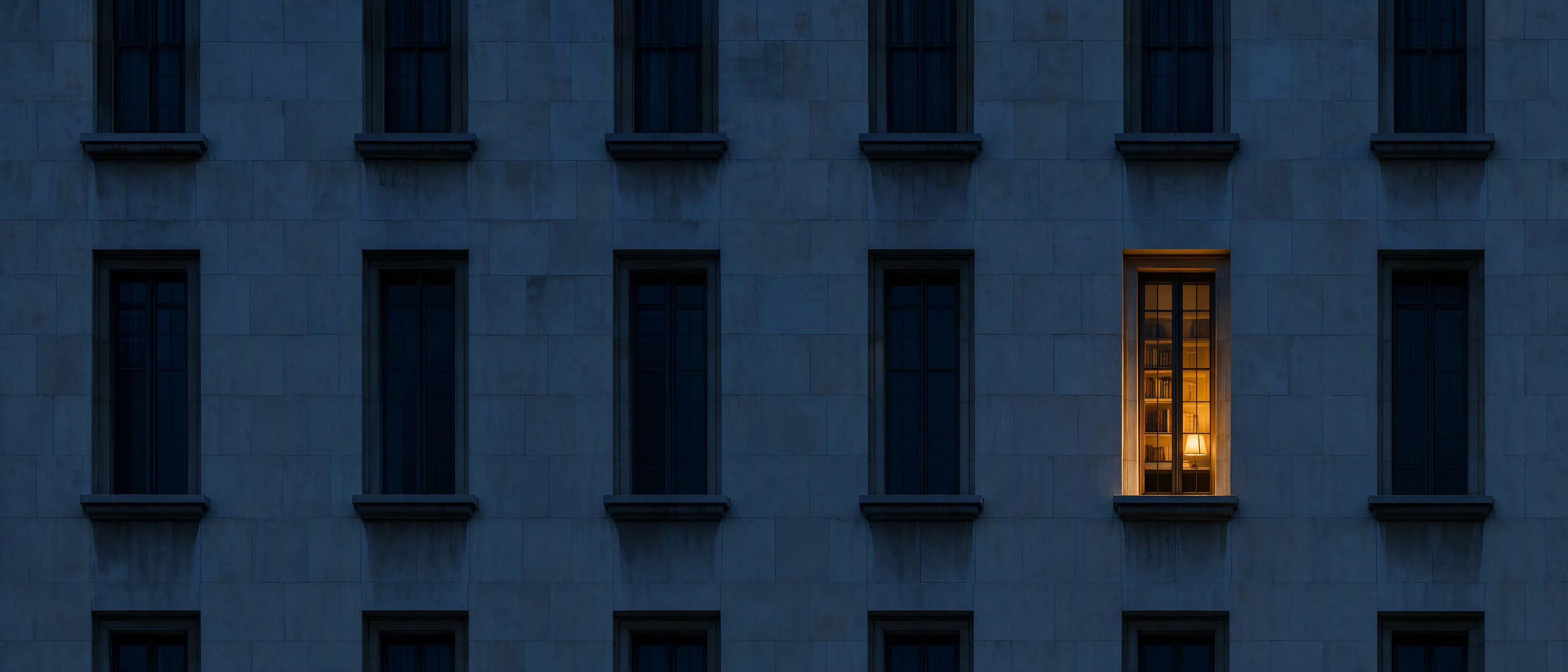 Dusk on the thick limestone wall of a government building, one deep-set window lit warm from inside.