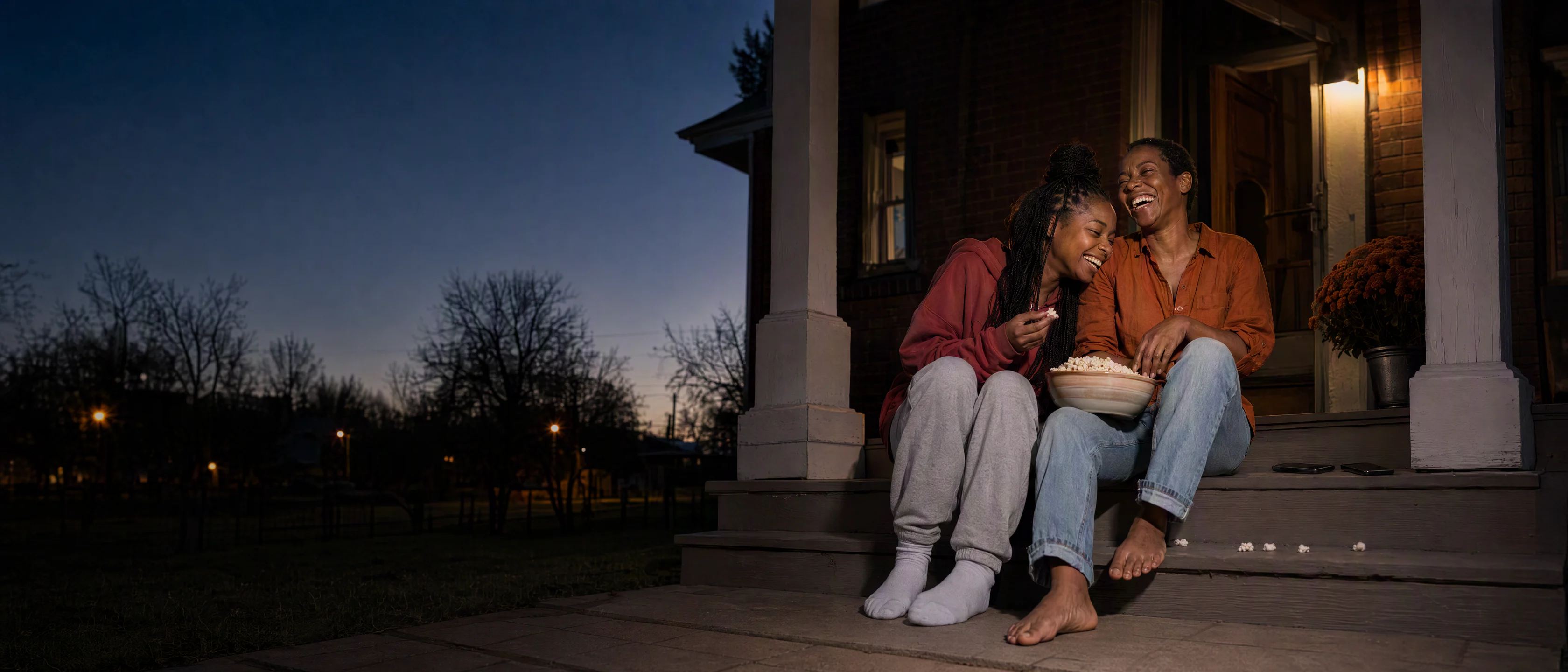 A mother and her college-age daughter laughing on their porch steps at dusk over a bowl of popcorn, their phones face down on the step beside them.