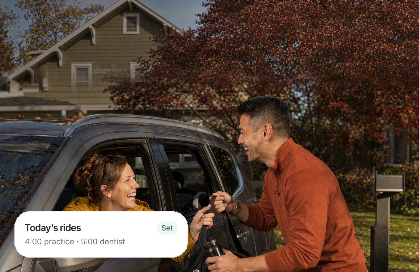 A dad at the curb handing his son’s soccer bag through a neighbor’s car window, both laughing; his Dvina card: today’s rides, practice at 4:00 and the dentist at 5:00.